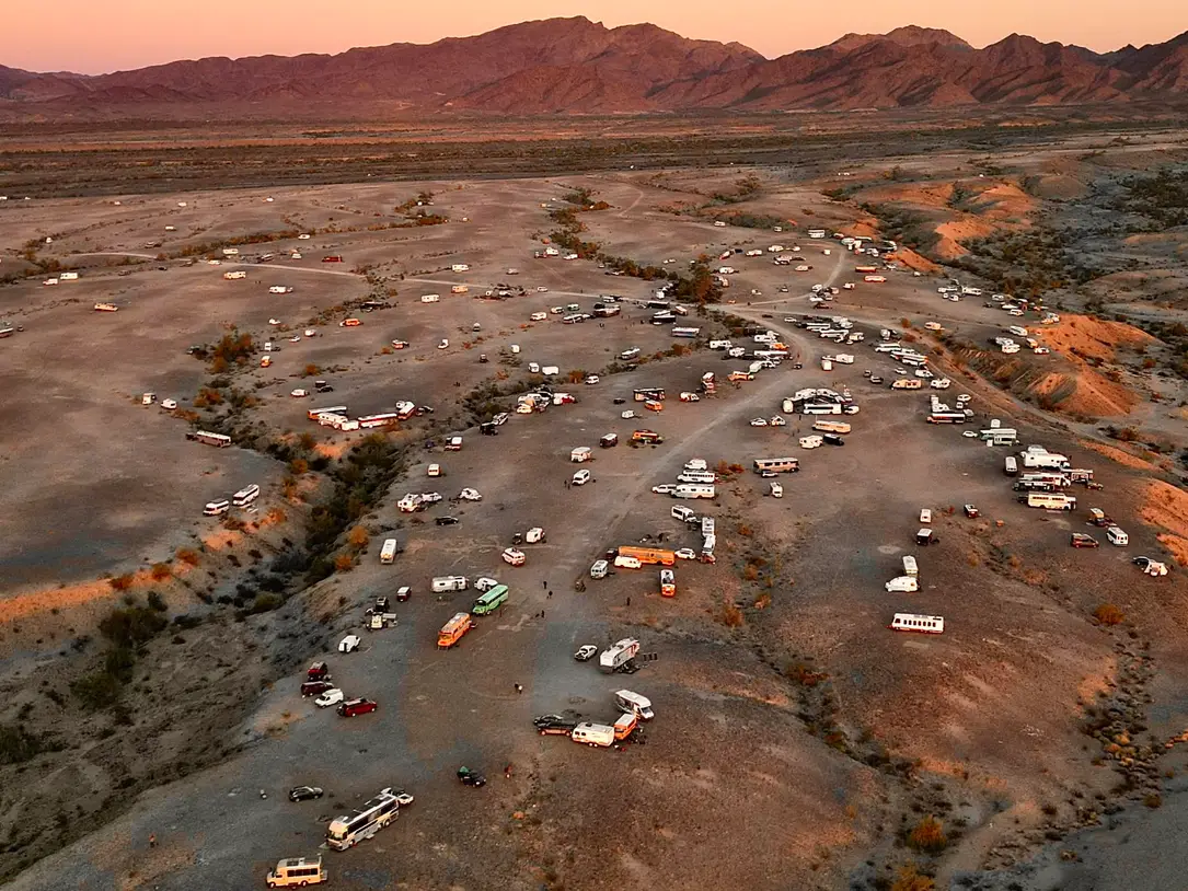 This meme is a screenshot of a popular social media platform's "Meme of the Year" award, featuring a picture of a desert festival with many RVs and cars in the foreground, with a large crowd of people in the distance....