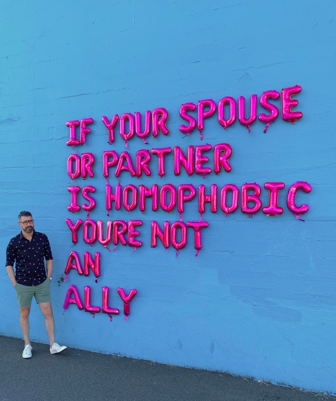 This meme is a photo of a man standing in front of a wall with a quote written in bold, pink, dripping letters. The quote reads: "If your spouse or partner is homophobic, you're not an ally. If your friend is racist, you're not an ally. If your family member is sexist, you're not an ally. If your colleague is transphobic, you're not an ally. If you're not actively working to dismantle the systems of oppression, you're part of the problem." The image is meant to be humorous and satirical, but also thought-provoking and provocative.