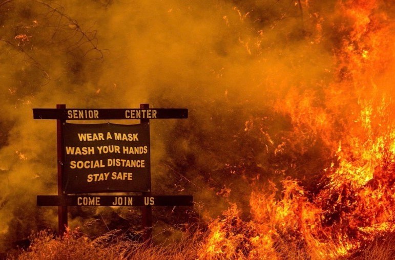 The image depicts a sign in front of a massive fire, with the sign reading "SENIOR CENTER" at the top and "WEAR A MASK, WASH YOUR HANDS, SOCIAL DISTANCE, STAY SAFE" below. The sign is surrounded by flames, indicating that it is located in a area where a fire is taking place.  The image is likely intended to be humorous, as it appears to be a situation where someone would expect to see a sign about safety and social distancing, but instead, it is surrounded by a massive fire. The image is likely meant to be seen as humorous or satirical, rather than serious.