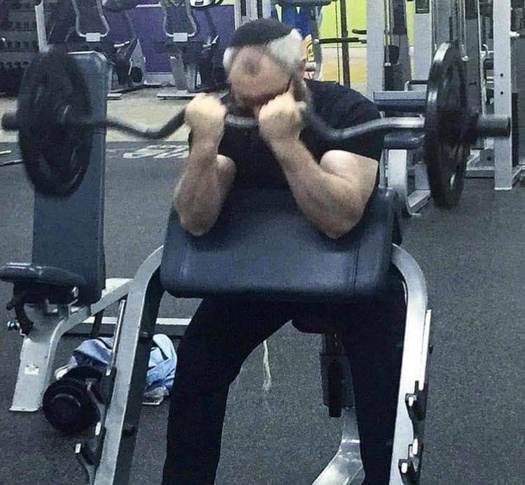 The image shows an older man with white hair and a black shirt working out at the gym. He is sitting on a weight machine, holding a barbell with both hands, and his face is pressed into the bar. His arms are bent and his hands are clenched, and his face is red and strained. The background is a gym with various machines and weights. The man is exerting himself to lift the weight, and his face is red and strained from the effort. The image is meant to be humorous, as it shows an older man trying to lift a weight that is likely too heavy for him, and his face is red and strained from the effort. The image is meant to be humorous, as it shows an older man trying to lift a weight that is likely too heavy for him, and his face is red and strained from the effort.