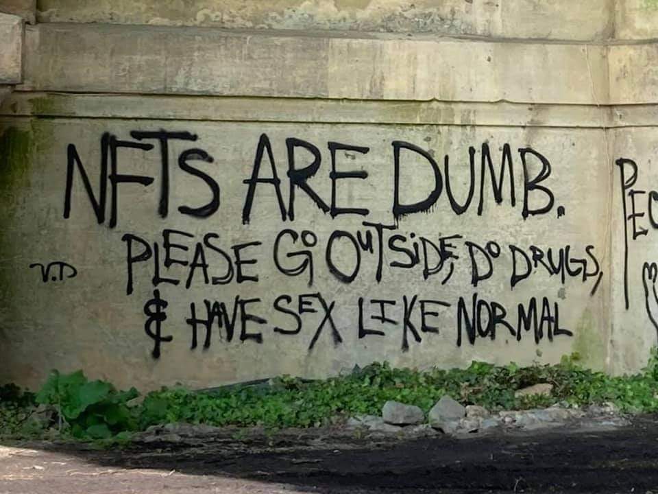 This meme is an image of a graffiti-covered wall with a handwritten note that reads: "NFTs are dumb. I'd rather have a root canal without anesthesia than buy one of these digital art pieces. Please go outside, do drugs, and have sex like normal people." The text is written in a sloppy, childish font, adding to the humorous and sarcastic tone of the message.
