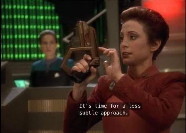 The meme features a still image from the TV show "Star Trek: The Next Generation." The image shows a woman holding a small, brown, triangular device and pointing at it. The text overlay reads, "It's time for a less subtle approach." The woman is likely Captain Jean-Luc Picard's chief of operations, Commander Deanna Troi, using her "empathic" device to scan the emotions of someone or something. The device is used to gather information about people and situations, and the woman is suggesting that they need to be more direct and open in their approach. The image is humorous because it implies that the woman is using her device to scan someone's emotions and is not getting the information she needs, so she's suggesting that they need to be more direct and open. The image is likely being used to poke fun at someone who is trying to be too subtle or indirect in their approach.