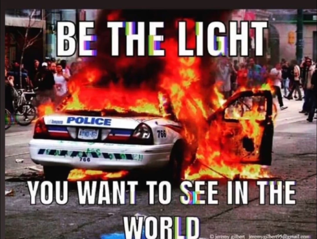 The meme features a car engulfed in flames with a police car in the background. The text reads: "BE THE LIGHT YOU WANT TO SEE IN THE WORLD." The image is meant to be ironic, as the car is on fire and the police car is being burned. The text is meant to be inspirational, but it is presented in a way that is opposite of its intended message. The image is meant to be humorous and satirical.