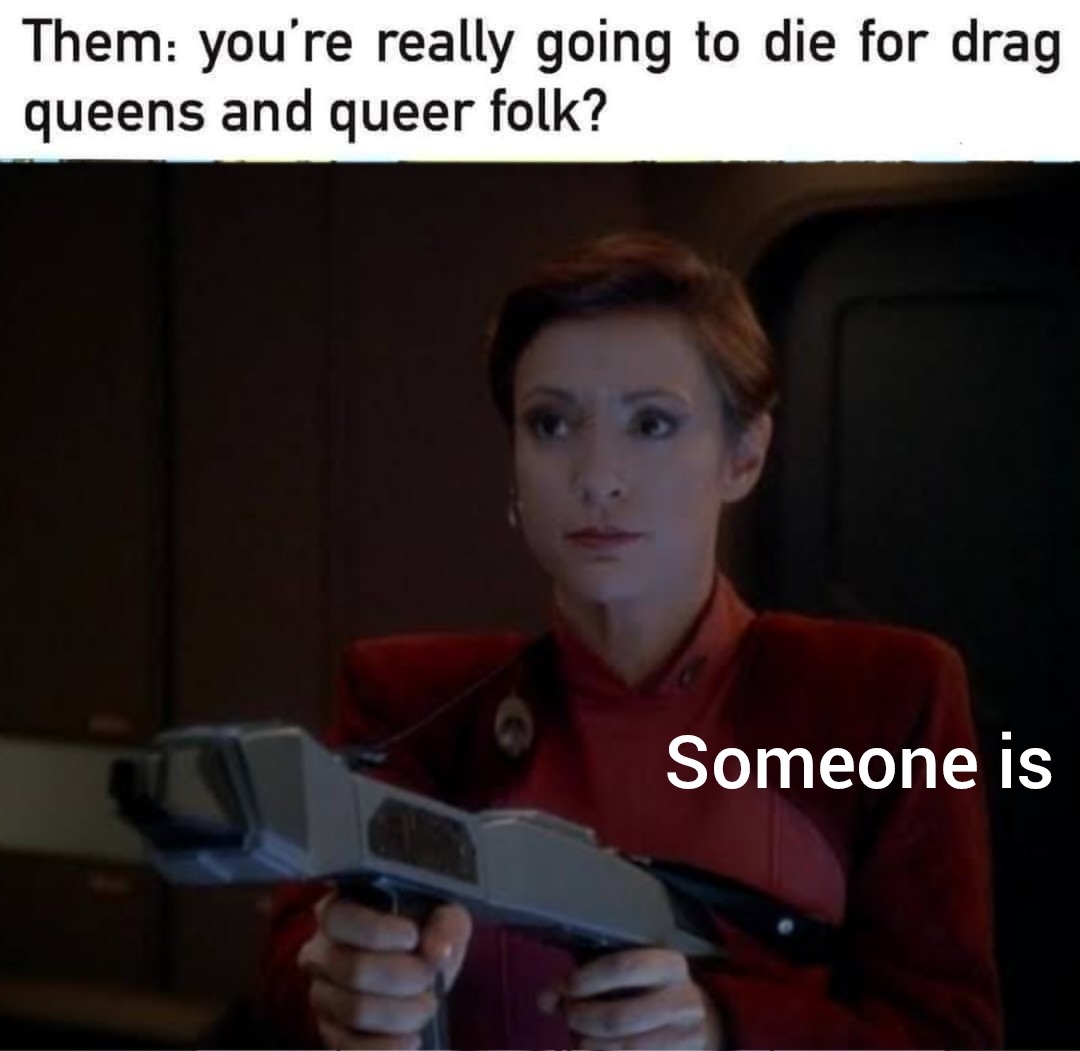 This meme is a humorous and unexpected response to a question about someone's death. The image shows a character from the Star Trek franchise, Captain Janis – who is holding a gun and looking at the camera with a serious expression. The text "Someone is" is overlaid on the image. The meme is a play on the idea of a person being asked to confirm their own death, and the response is a deadpan "Someone is" – implying that someone is indeed dead.