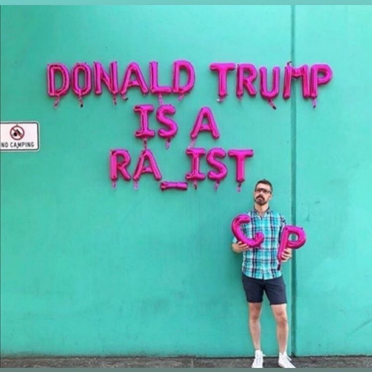 This meme features a man holding a sign that says "Donald Trump is a racist" in a childlike, blocky font, but with a key typo: "racist" is spelled "racist" with a "c" replaced by a "c" with a diagonal line through it, indicating the word "c" is actually "c" with a line through it, or "c" with a "c" replaced by "c".
