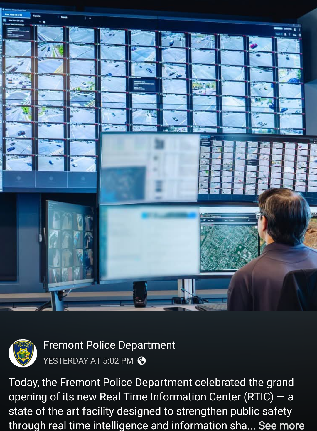 This photo depicts a man sitting in a control room facing an array of monitors filled with grid-based video feeds, likely monitoring traffic and intersections. To the right, one screen displays green dots on a map, wh...