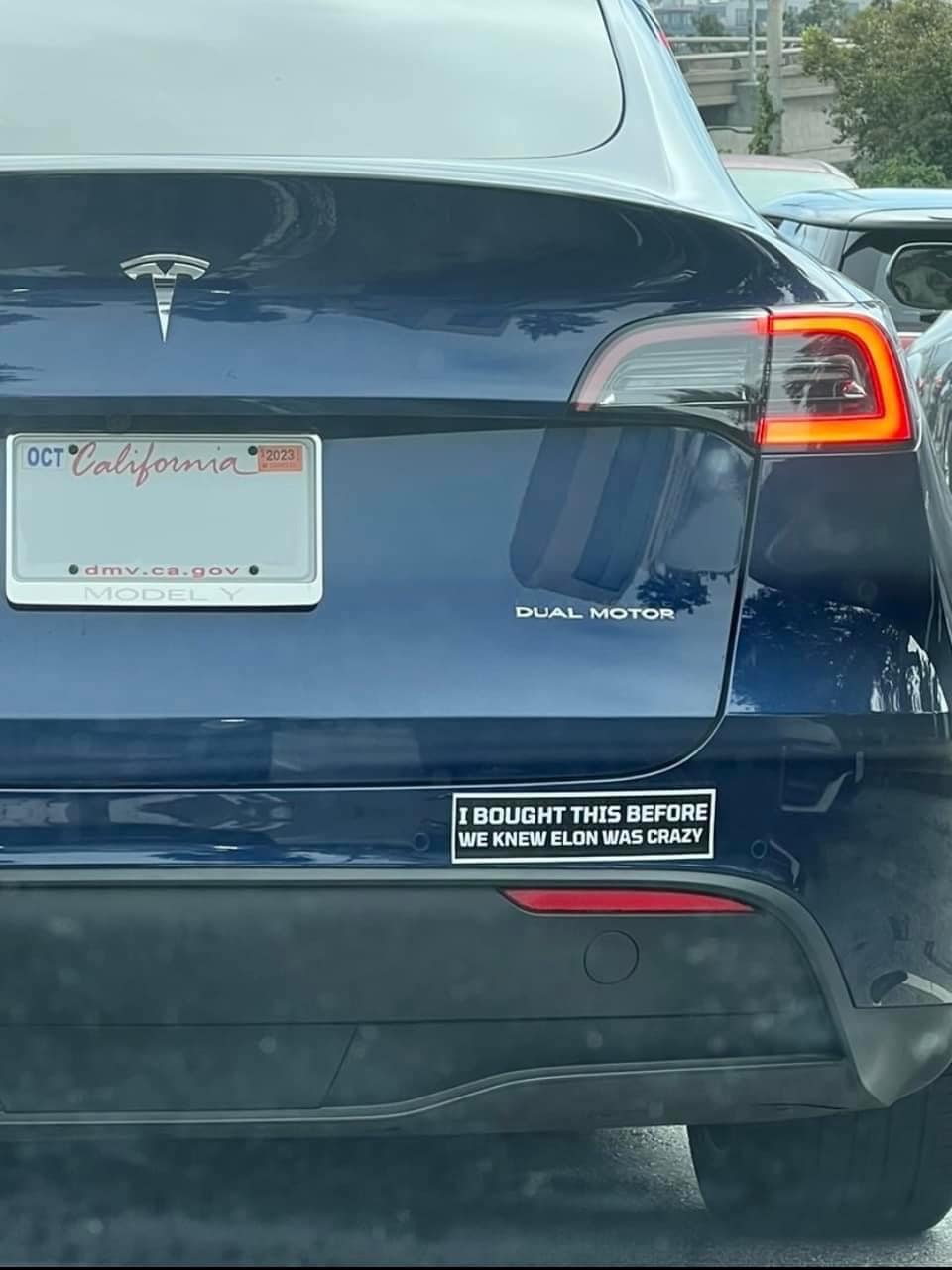 The meme is a humorous take on the popular "I'm not a [insert profession] I'm a [insert profession]" joke. The image shows the back of a Tesla with a bumper sticker that reads "I bought this before we knew Elon was crazy". The joke is a play on the popular meme format, but with a Tesla-specific twist, referencing the eccentric billionaire's reputation.