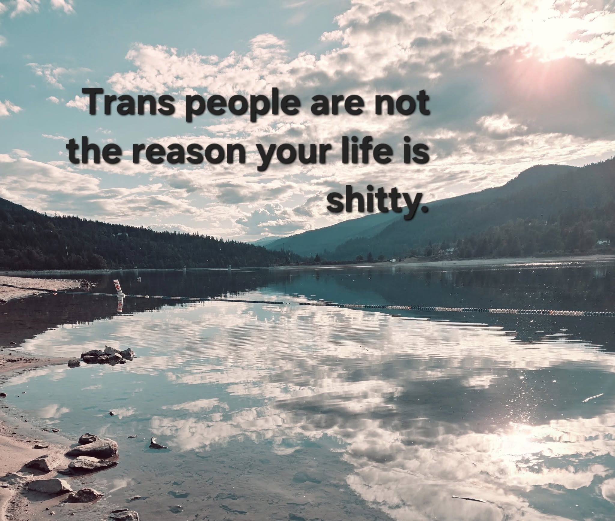 This meme is a photo of a serene lake surrounded by mountains, but the image is intentionally distorted and has a "vintage" or "glitchy" aesthetic. The text "Trans people are not the reason your life is shitty" is overlaid on top of the image, with a bold and straightforward font. The text is meant to be a humorous and sarcastic response to people who blame trans individuals for their own problems.