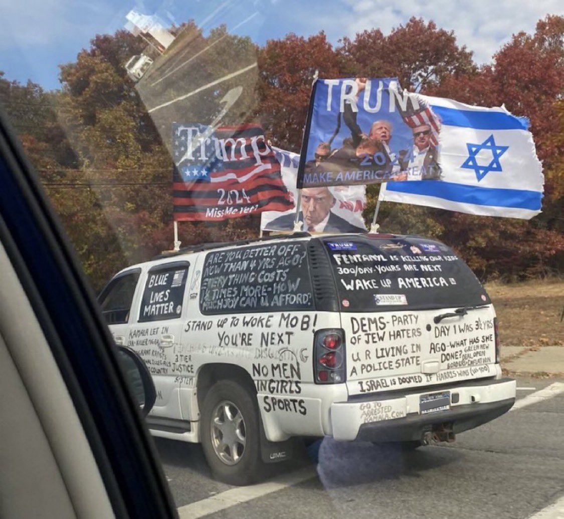 This meme is a photo of a car with a heavily modified and decorated exterior, featuring a mix of pro-Trump and anti-Trump messages, as well as some anti-Semitic and racist graffiti. The car's windows are covered in scribbled notes, stickers, and flags, with phrases like "Make America Great Again" and "God is Trump" alongside more inflammatory and hateful language. The car's bumper reads "I [heart] Trump" and "God is Trump".