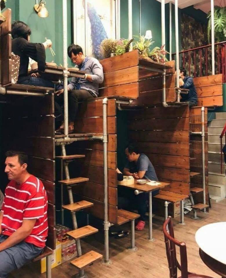 The meme is an image of a crowded restaurant with people sitting on tables, stairs, and even the floor, with others sitting on top of tables and even on the ceiling. The image is captioned "When you're trying to get a table at a popular restaurant and you're still waiting". The meme pokes fun at the frustration of waiting for a table at a busy restaurant, exaggerating the situation to absurdity.