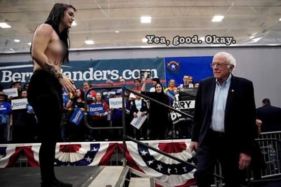 This meme is a still image of a 2020 US presidential campaign event. Bernie Sanders, a white-haired, elderly man with a long, white beard, is standing in front of a crowd of people. He is wearing a suit and looking at a young, topless woman with long, dark hair, who is standing on a table. The text "Yea, good. Okay." appears above the image, suggesting that the woman's presence is not causing any concern or reaction from Sanders.