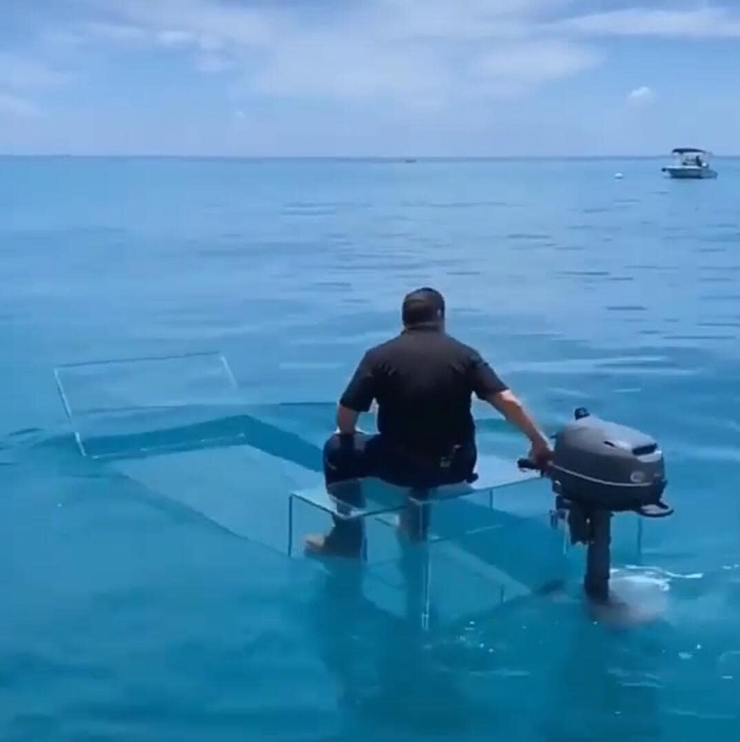 The meme depicts a man sitting on a transparent glass table in the middle of the ocean, with a small motorboat nearby. The image is captioned "When you finally get that promotion and get to work from home". The humor lies in the absurdity of the situation, as the man is literally "working from home" in a literal sense, surrounded by water.