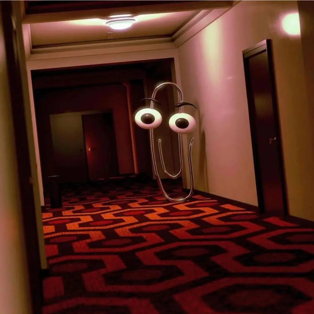 This meme features a screenshot from the 1984 film "The Shining" with a character from the movie's hotel environment, but the character is replaced with the computer icon from the "Clippy" software that was used in the 1990s. The image is captioned "Clippy just got a job at the Overlook Hotel".