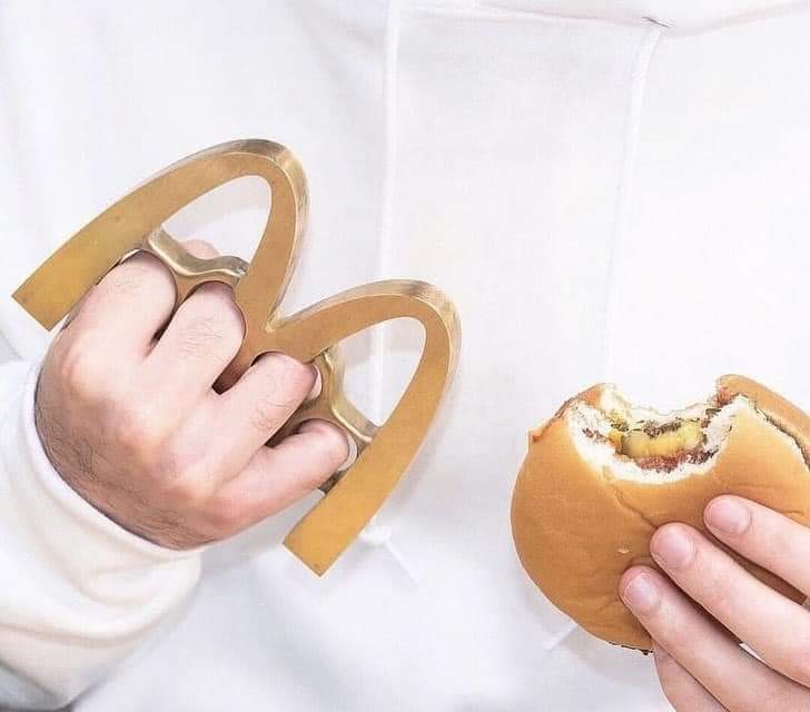 The meme is a humorous image of a person holding two large, metal "fingerless" gloves, also known as brass knuckles, while eating a burger. The text "I'm not arguing, I'm just explaining why I'm eating my burger with brass knuckles" is often added to the image, implying that the person is being aggressive or confrontational while eating. The meme is a playful way to poke fun at someone who is being overly assertive or argumentative.