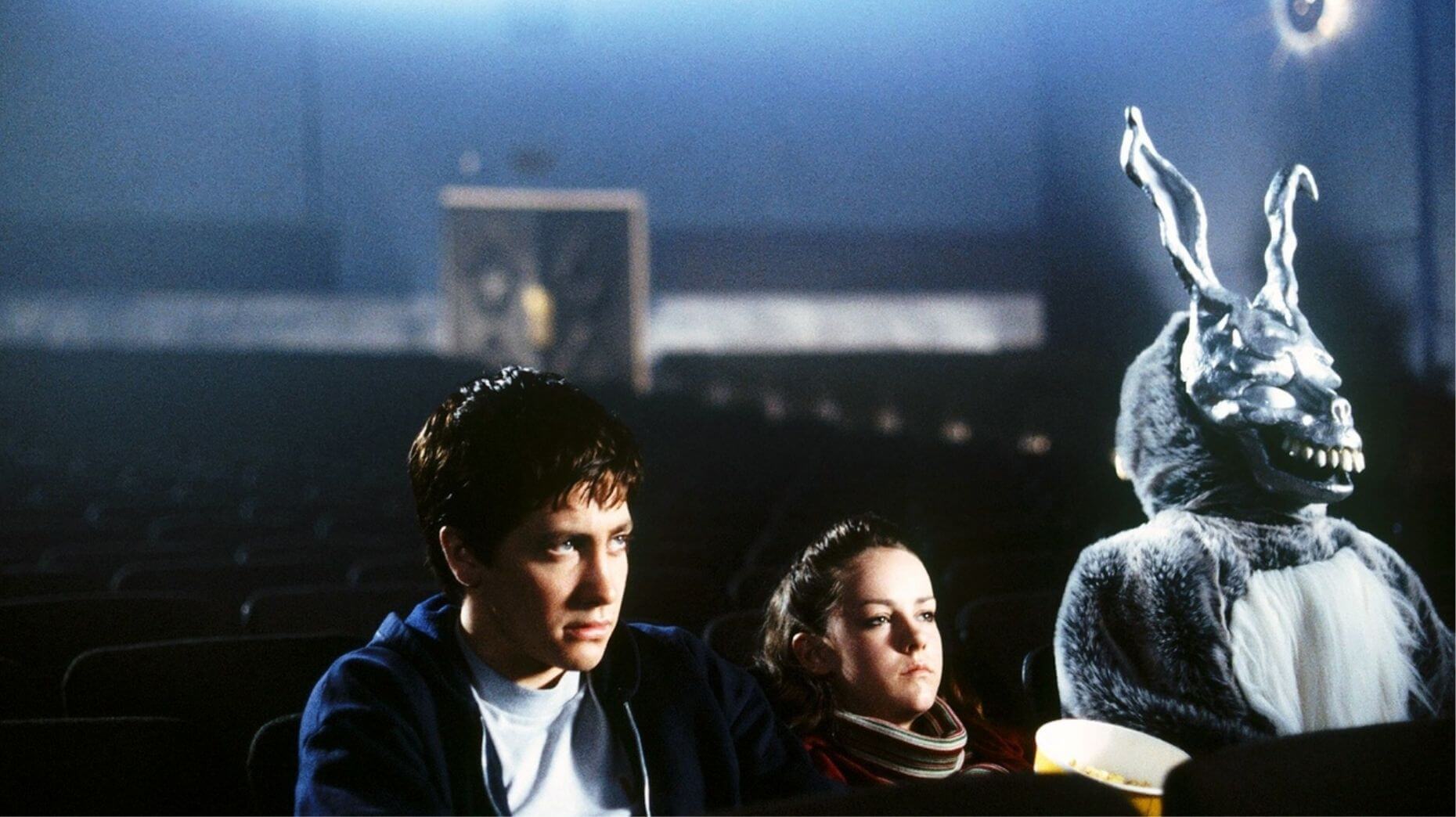 This meme is a screenshot from the movie "Donnie Darko" featuring the main characters Donnie and his girlfriend Elizabeth. The image shows a dark and eerie atmosphere, with Donnie and Elizabeth sitting in a dimly lit theater, surrounded by an unsettling presence. The text overlayed on the image reads "When you're trying to watch a movie but the existential dread of the universe is just sitting in the seat next to you".