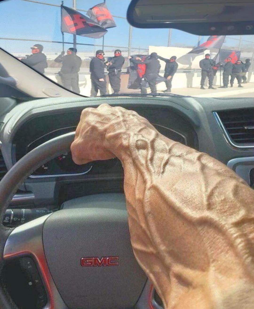 The meme is a photo of a person's arm, likely a bodybuilder or fitness enthusiast, with a large, muscular arm flexed and resting on the steering wheel of a car. The image is captioned "Driving through a protest".
