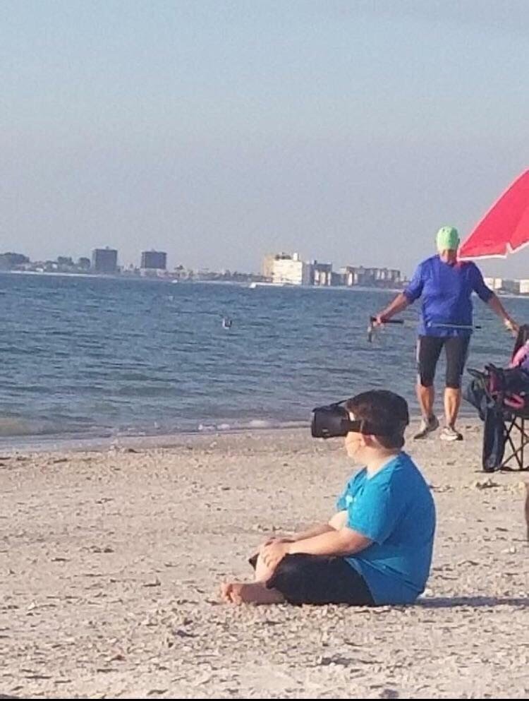 This meme is an image of a person sitting on a beach, wearing a VR headset and seemingly oblivious to their surroundings. The image is captioned "I'm not ignoring you, I'm just in a different world". The meme is likely humorous because it pokes fun at the idea of using VR to escape reality, but in this case, the person is literally escaping their surroundings on a beach.