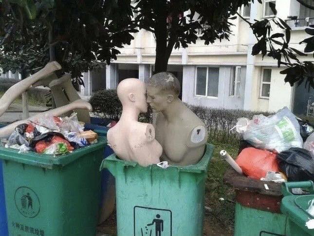 The meme features two mannequins in a romantic pose, leaning in to kiss each other, in a trash-filled alleyway. The image is captioned "I love you, even in the trash". The juxtaposition of the romantic gesture with the trash-filled setting creates a humorous and ironic contrast.