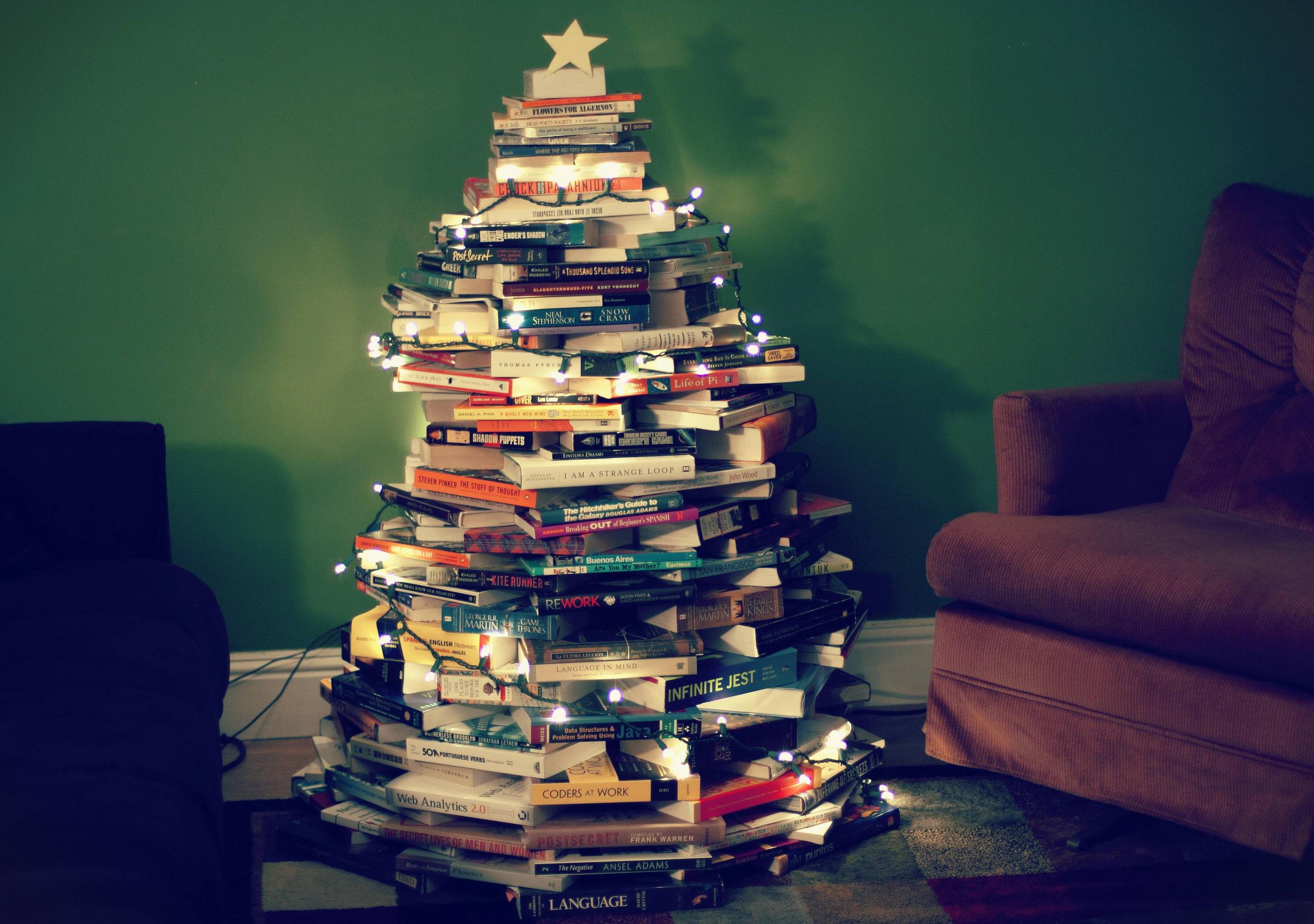 The meme is an image of a Christmas tree made entirely out of books, with strings of lights and a star on top. The tree is surrounded by a cozy living room, with a couch and a coffee table in the background. The image is captioned "I've been reading a lot this year".