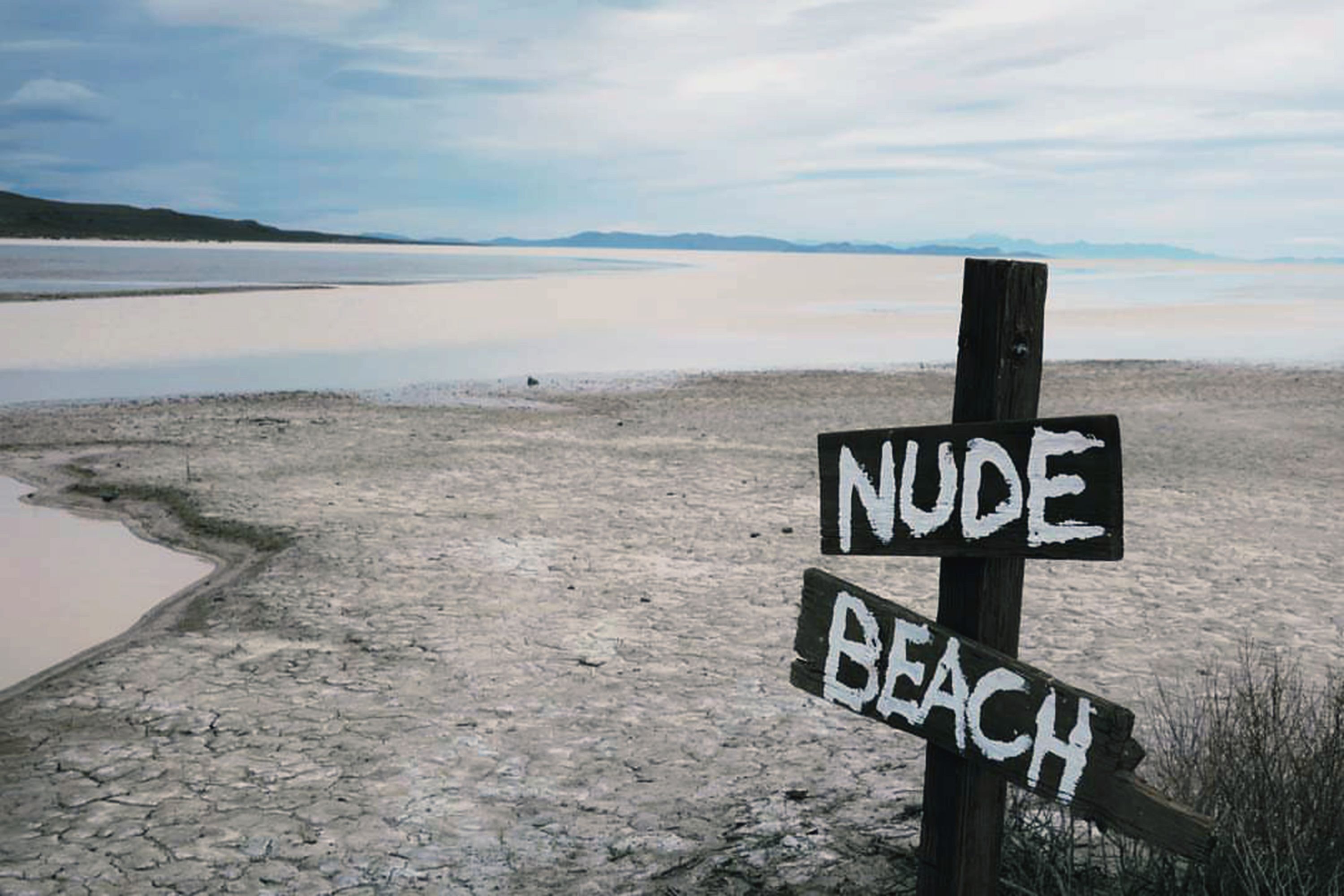 The meme is a humorous image of a sign that says "Nudist Beach" with an arrow pointing to the left, but the sign is actually pointing to a dry, cracked lake bed with no water in sight. The text "Nudist Beach" is written in a playful, hand-drawn font, but the image is a stark contrast to the usual expectation of a beach. The joke is that the sign is pointing to a desolate, barren landscape instead of a beautiful beach.
