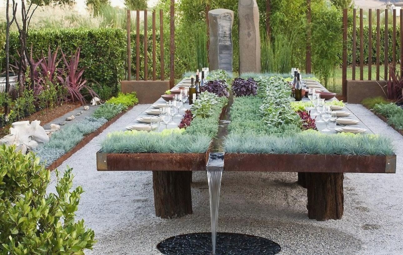 The meme is a photo of a serene garden with a large stone table and chairs, surrounded by lush greenery and a small pond. The caption reads "When you finally get a garden that's perfect for dinner parties, but the guests are all plants".