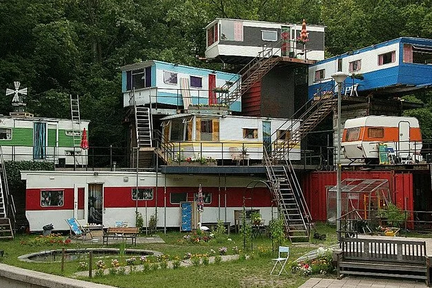 The meme is an image of a group of colorful, small houses stacked on top of each other, with a garden and a few people sitting outside. The text "Permaculture - Shelter" appears at the top of the image, suggesting tha...