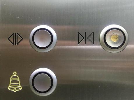 This meme is a screenshot of the "Dumb" button on a elevator control panel. The button is highlighted in yellow and has a red circle around it, indicating it has been pressed. The text "Dumb" is written above the button in a bold font.