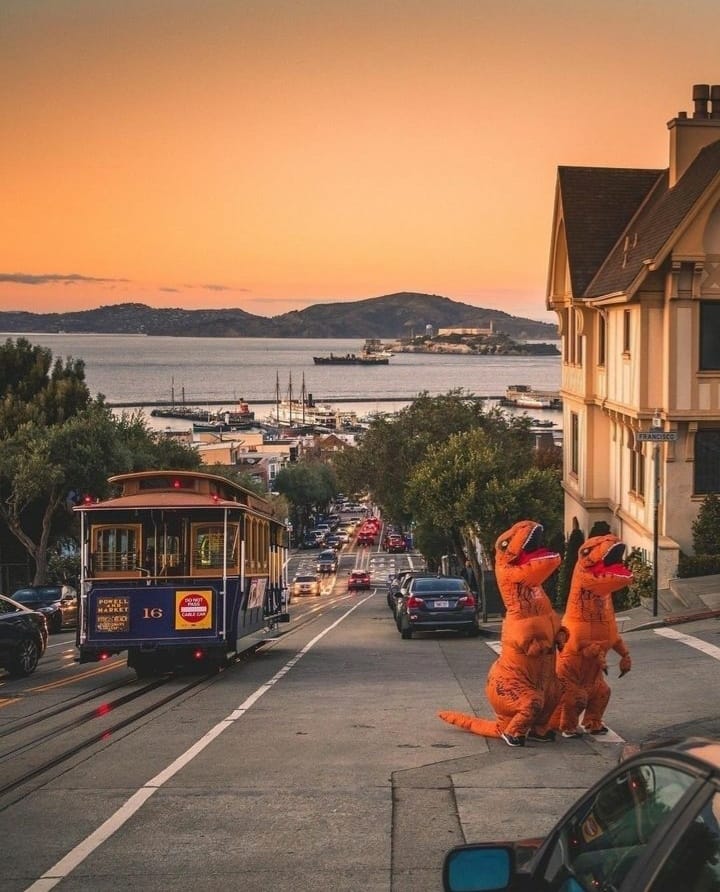 The meme is an image of two people in full-body T-Rex costumes standing on a San Francisco cable car track, looking out at the city's iconic Golden Gate Bridge. The image is captioned "When you're trying to get to the other side but you're stuck in traffic".