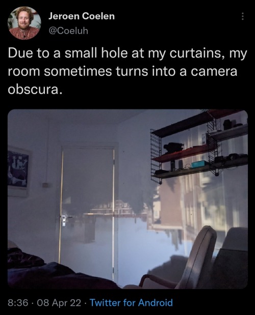 The meme is a screenshot of a Twitter post with a photo of a dark room with a faint, eerie glow coming from a hole in the wall. The text overlayed on the image reads: "Due to a small hole in my wall, my room sometimes turns into a camera obscura."
